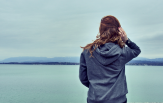 Woman standing alone looking out over calm teal water, reflecting the quiet and isolating experience of living with chronic pain.