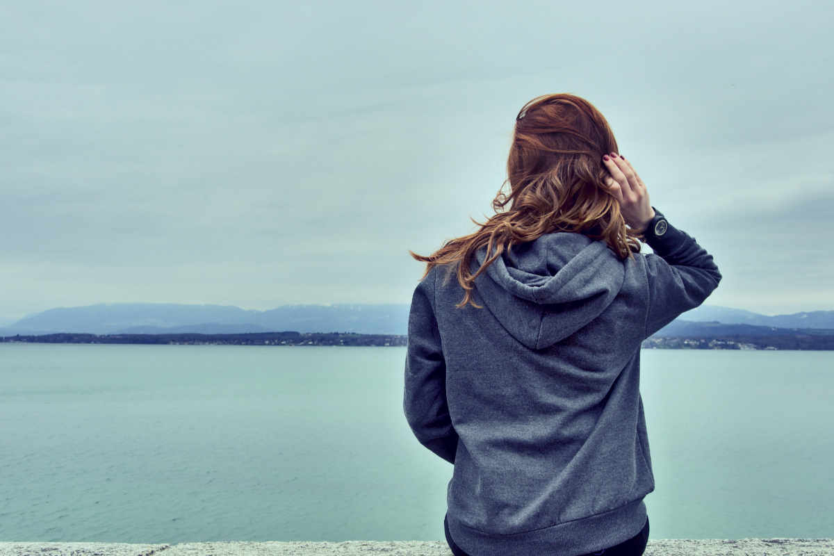 Woman standing alone looking out over calm teal water, reflecting the quiet and isolating experience of living with chronic pain.